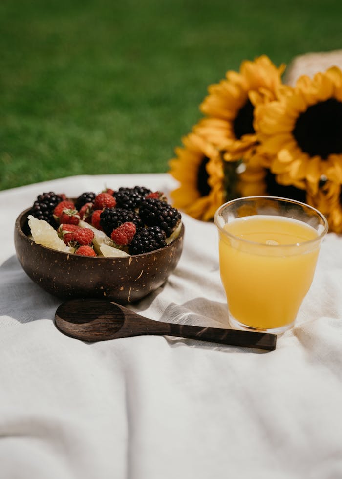 Fruit bowl with berries and orange juice on picnic blanket with sunflowers outdoors.
